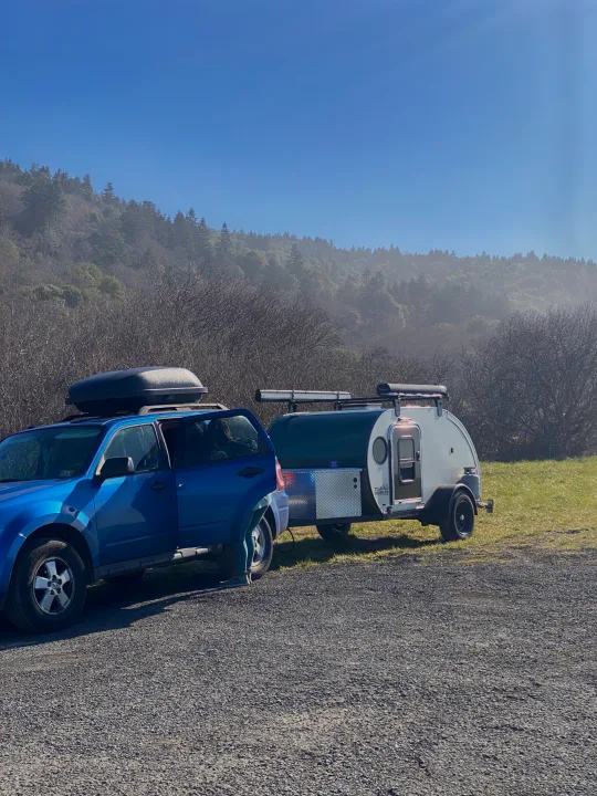 Blue SUV with roof box towing a compact teardrop RV rental. Parked in a gravel lot with misty forest background.. Teardrops Northwest Trek 2022