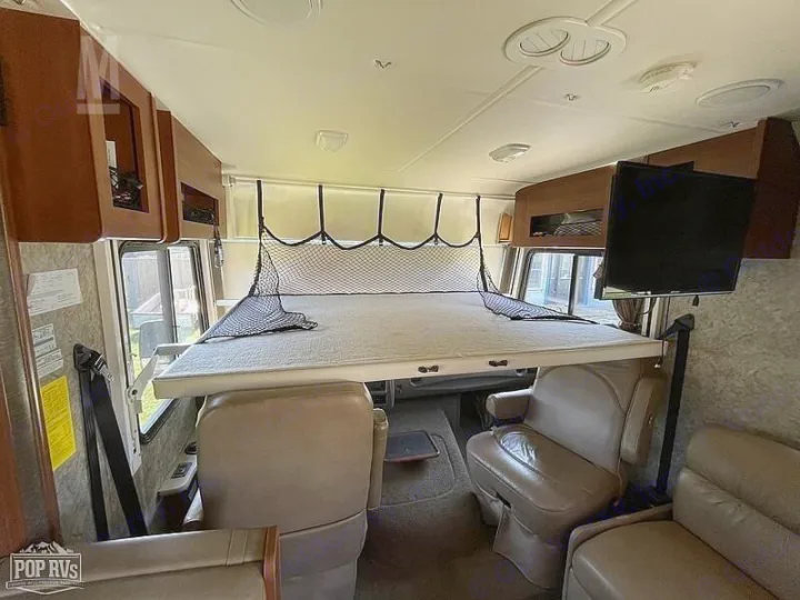 RV rental interior with a deployed bunk bed over front seats, featuring a TV, cabinets, and seating areas.. Fleetwood Other 2015