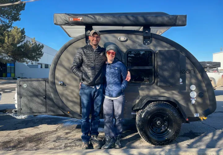 A happy couple poses with their sleek, dark grey teardrop camper, ideal for an RV rental adventure. Equipped with a 23ZERO awning.. Bean Mean Bean 2022