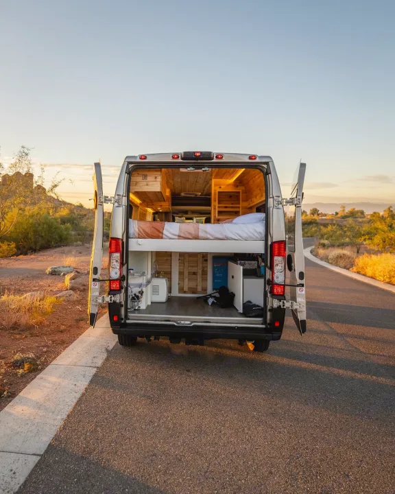 Rear doors open on an RV rental camper van, revealing its cozy bed, wood interior, and equipment for desert travel at sunset.. Dodge Ram Promaster 2500 High Roof 159" 2021