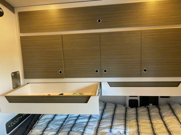 Inside an RV rental: Modern overhead storage cabinets, an open drawer, and striped bedding on a bed. Compact living for travel.. Bean Mean Bean 2022