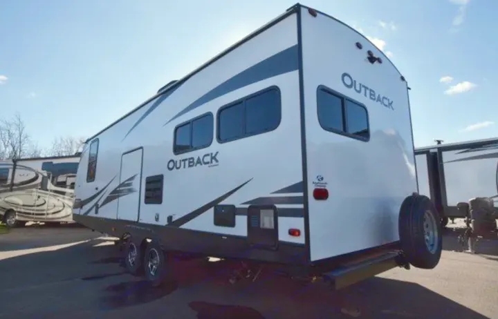 This white Outback travel trailer, an excellent RV rental option, is parked outdoors. It features multiple windows & a rear spare tire.. Keystone RV Outback Ultra-Lite 2020
