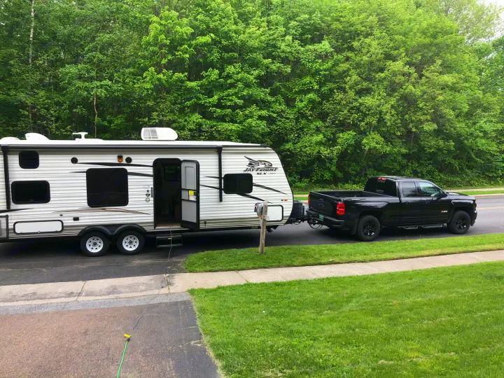 A white travel trailer RV rental, hitched to a black pickup truck, is set for adventure. Lush trees provide a natural backdrop.. Jayco Jay Flight SLX 2017