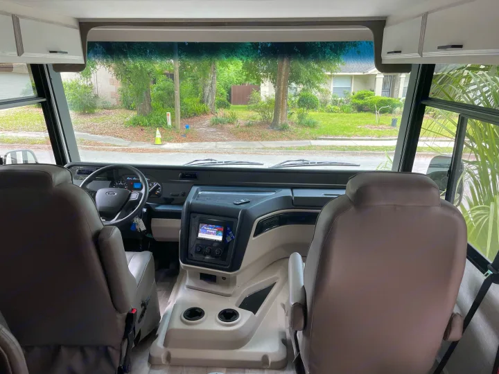 Inside an RV rental: driver's cockpit, dashboard, and steering wheel, with a clear view of trees and houses outside.. Other Other 2022