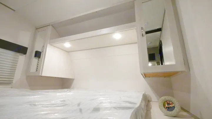 Modern RV rental bedroom. A bed in protective plastic sits under light-wood overhead cabinets with mirrored doors & integrated lights.. Prime Time RV 260BHSLE 2022