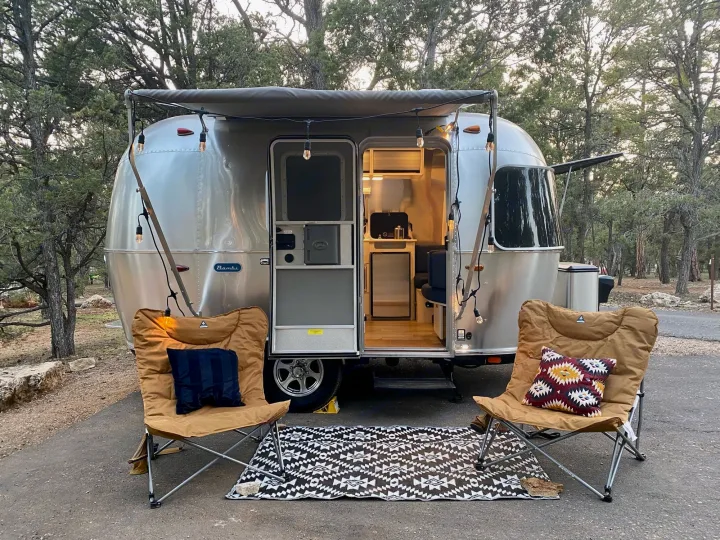 A silver Airstream Bambi rv rental is parked with an awning and string lights. Two chairs and a mat create a cozy outdoor setup.. Airstream Bambi 2021