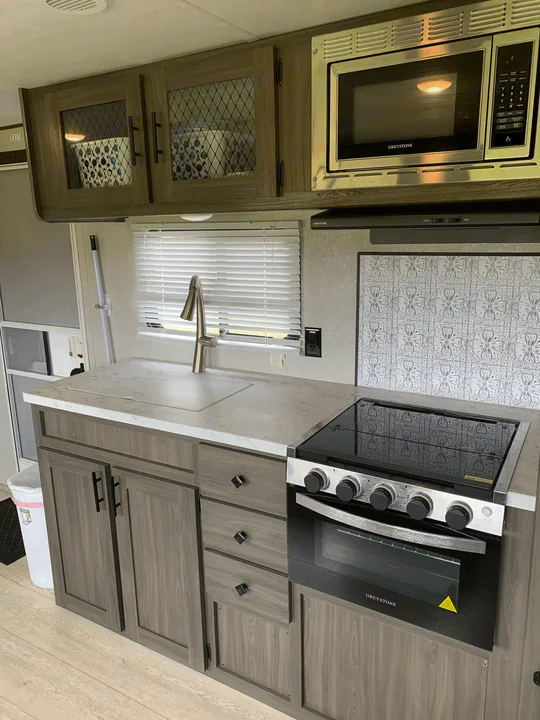 Well-appointed RV rental kitchen with modern light wood cabinets, Greystone microwave, cooktop, and sink.. Other Other 2021
