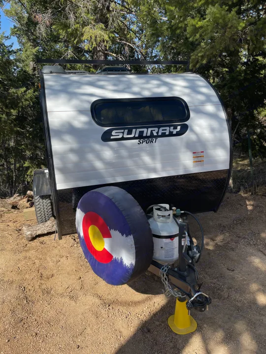 Compact Sunray Sport teardrop camper rv rental, featuring a Colorado flag tire cover & propane tank, nestled in the woods.. Sunset Park RV SunRay 2020
