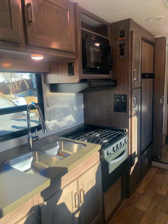 Modern RV rental kitchen with a double sink, gas stovetop, microwave, and refrigerator, all within wood-look cabinetry.. Winnebago Minnie Winnie 2020