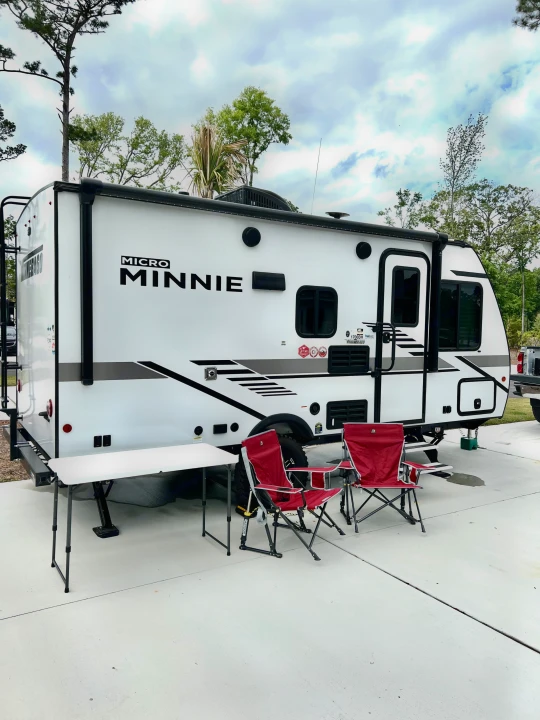 This Micro Minnie travel trailer RV rental, parked on a pad, offers comfortable camping with red chairs and a table set up outdoors.. Winnebago Micro Minnie 2021