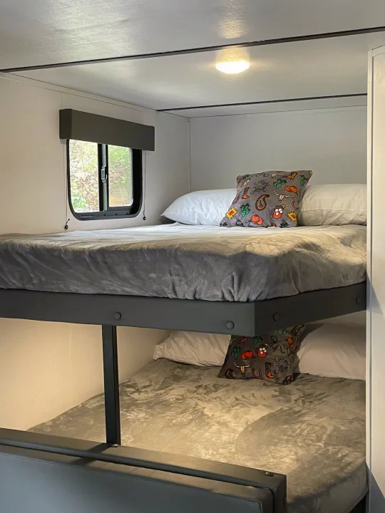 Cozy bunk beds in an RV rental with gray bedding, patterned pillow, window, and overhead light for a comfy stay.. Keystone Springdale 2022