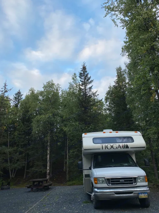 An RV rental parked at a serene forest campsite with a picnic table under a cloudy sky.. Fleetwood Tioga 2004