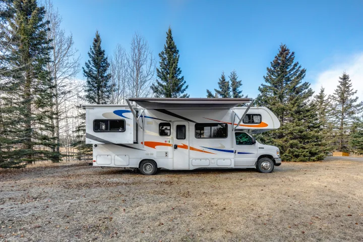 White Class C RV rental with awning extended, parked in nature among trees under a clear blue sky.. Forest River Sunseeker 2014
