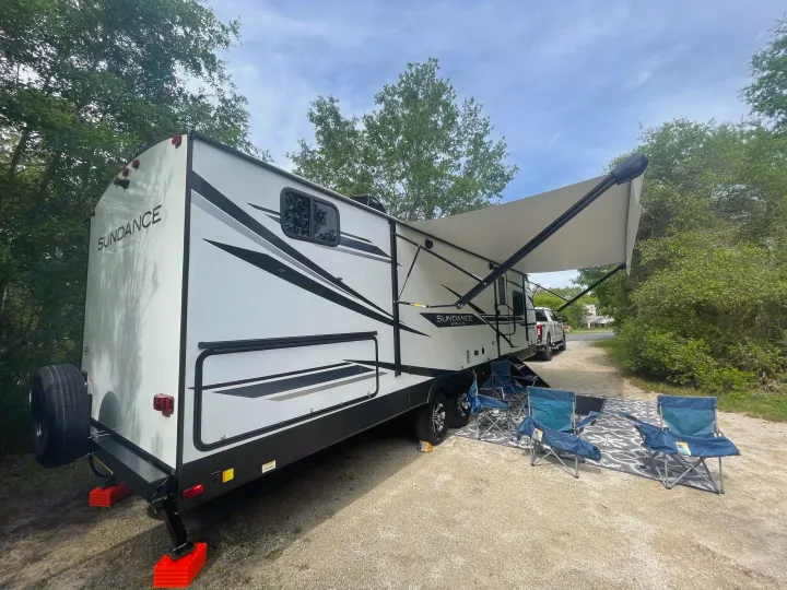 A white Sundance Ultra Lite rv rental is set up at a gravel campsite, complete with an extended awning, chairs, and a mat.. Heartland Sundance 294BH 2022