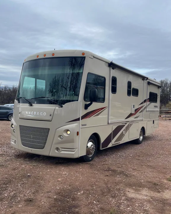 A beige Winnebago Vista RV is parked on a gravel lot under a cloudy sky, ready for your next rv rental.. Winnebago Vista 2018
