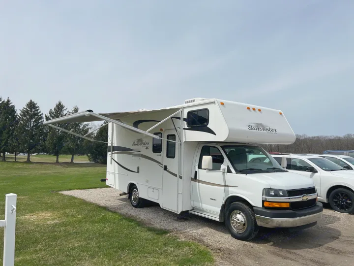 RV rental: White Sunseeker Class C RV with awning extended, parked on gravel beside green grass and distant trees.. Forest River Sunseeker LE 2013