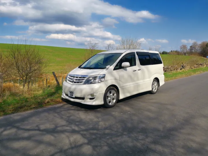 A white Toyota Alphard van, suitable for an RV rental, parked on a sunny country road with green fields under a cloudy sky.. Toyota Other 2022
