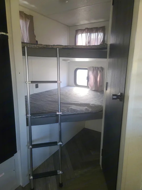 RV rental interior view: a compact sleeping area with bunk beds, a ladder, and a sunlit window.. Forest River Wildwood FSX Platinum 2021