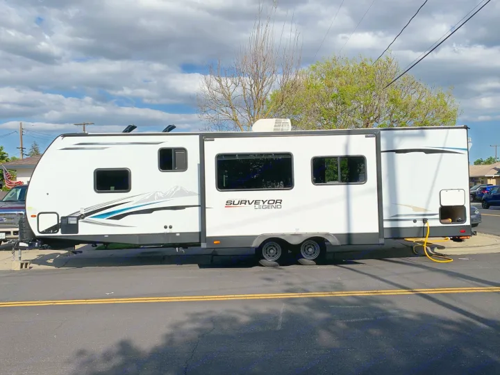 A large, white rv rental travel trailer is parked on a sunny street. Its side slide-out is extended, offering ample space.. Forest River Surveyor Legend 296QBLE 2021