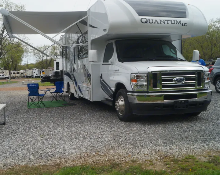 White RV rental, a Quantum LC motorhome on a gravel lot with extended awning & blue chairs for outdoor camping relaxation.. Thor Quantum 2022