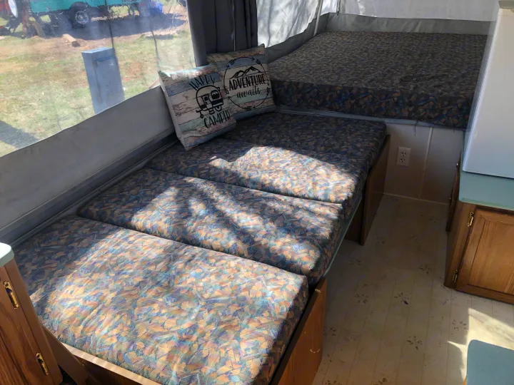 Interior of an RV rental featuring patterned bench cushions, decorative 'Happy Camper' pillows, and a sunlit window view.. Coleman Chesapeake 1993