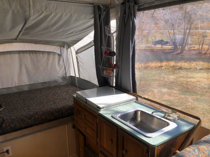 RV rental interior: cozy pop-up camper with a bed, small kitchen sink, and a scenic window view of trees and nature.. Coleman Chesapeake 1993