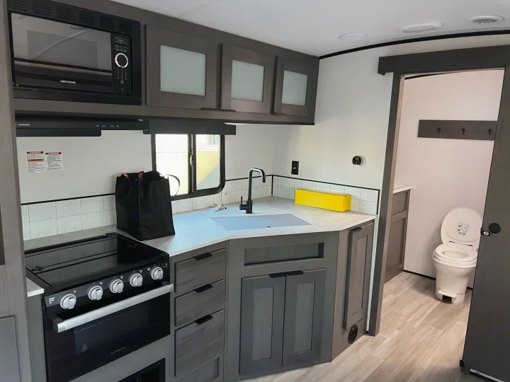 Modern RV rental kitchen featuring microwave, stovetop, sink, and a peek into the bathroom with a toilet.. Other Other 2021