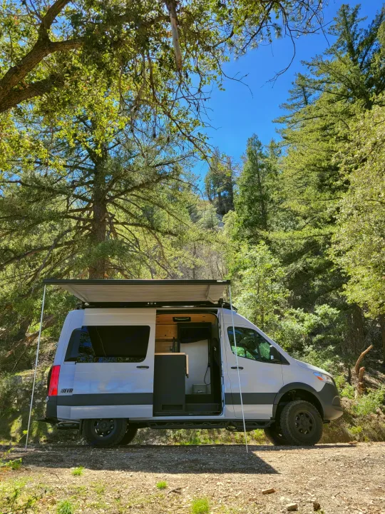 White camper van with open door and extended awning parked on a forest dirt road under blue sky. Perfect for your next RV rental adventure.. Mercedes-Benz Sprinter 2021