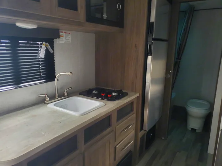This RV rental interior showcases a compact galley kitchen with a sink, stovetop, microwave, and fridge, plus a private bathroom with a toilet.. Crossroads RV Zinger 2021