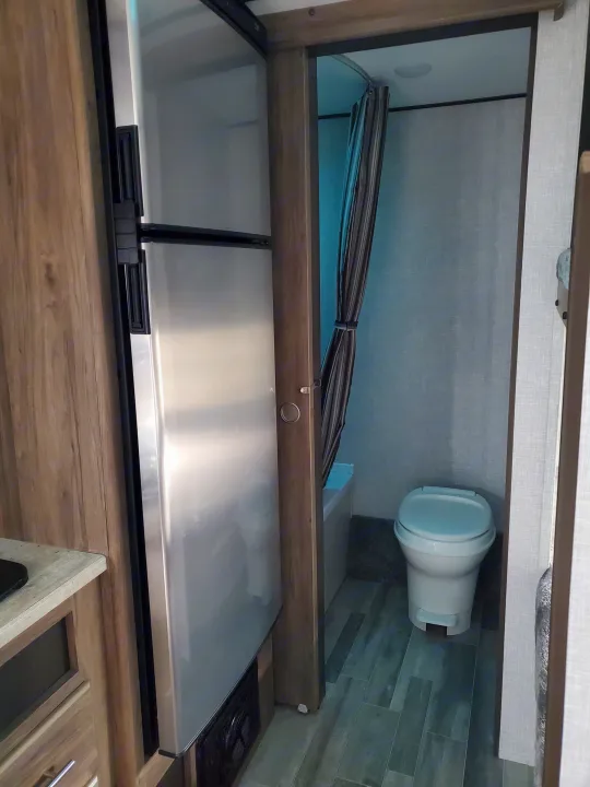 RV rental interior showing a stainless steel refrigerator and a compact bathroom with a toilet and shower curtain.. Crossroads RV Zinger 2021