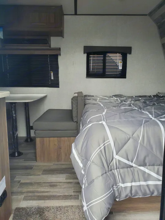 Cozy RV rental interior featuring a gray comforter on a bed, adjacent dinette area, and dark wood overhead cabinets.. Crossroads RV Zinger 2021
