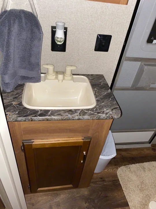 This RV rental bathroom offers a compact cream sink with a dark countertop, wooden cabinet, and wall-mounted air freshener.. Keystone RV Springdale 2017