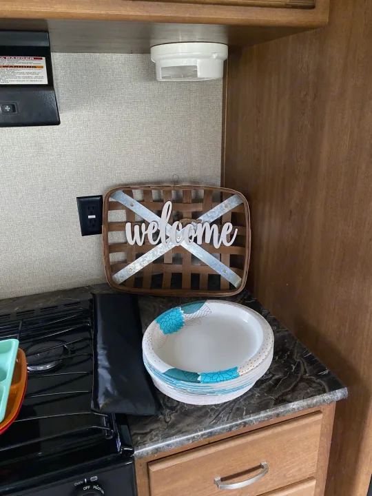 Inside an RV rental: compact kitchen with stovetop, "welcome" sign, paper plates, and essential safety features.. Keystone RV Springdale 2017
