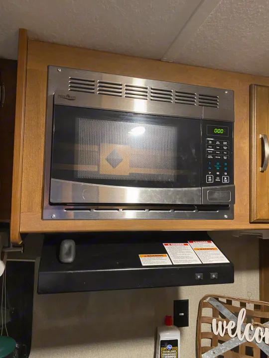 High Pointe microwave and range hood in an RV rental kitchen. Features a compact design, warning labels, and wooden cabinetry.. Keystone RV Springdale 2017