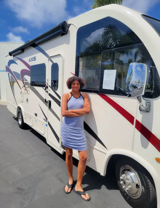 Young woman with a happy smile poses confidently beside her Axis RV rental, ready for a travel adventure.. Thor Axis 2021