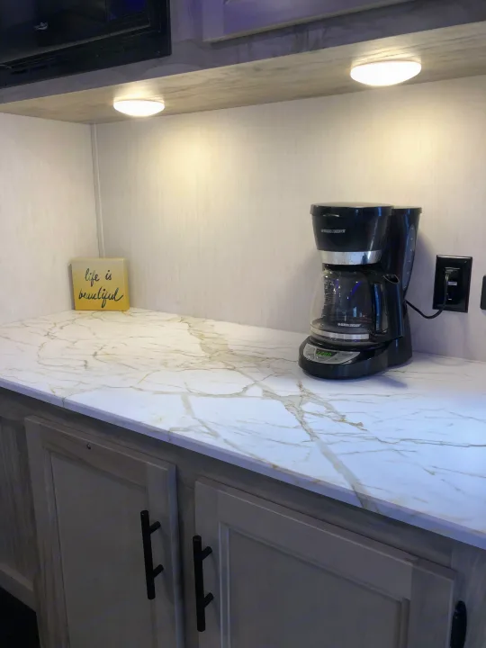 Modern RV rental kitchen counter with marble-look surface, black coffee maker, and under-cabinet lighting. "Life is beautiful" sign.. Forest River Cherokee Alpha Wolf 2022