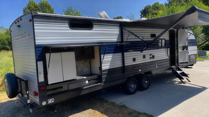 Sunny day view of an rv rental: a silver and blue travel trailer with its outdoor kitchen open, awning extended, and entry steps down.. Forest River Cherokee Grey Wolf 2020