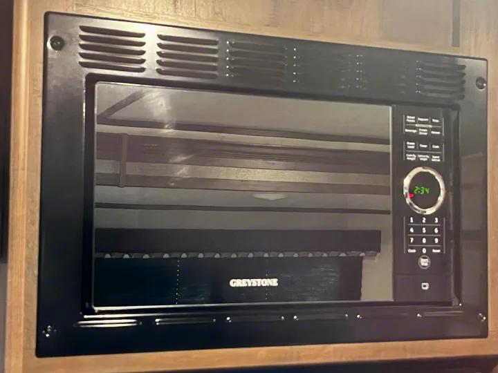 Built-in black microwave oven with "2:34" on display, reflecting an RV interior with "GREYSTONE" blinds. Perfect for an RV rental.. Coleman QBWE295 2021