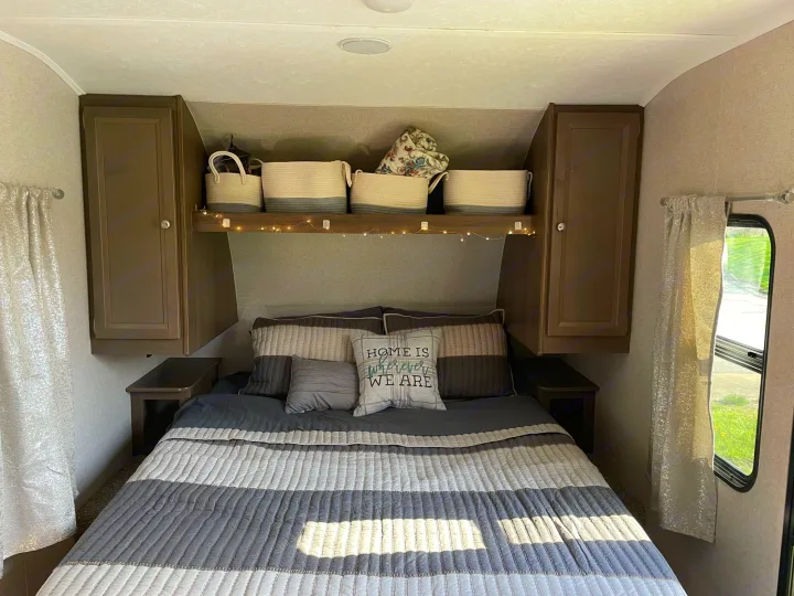This inviting RV rental bedroom offers comfort with striped bedding, ample overhead storage, and quaint decor.. Forest River Independence Trail 2021