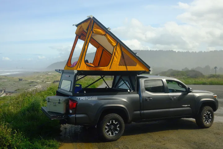 A dark grey Toyota Tacoma with an orange rooftop tent deployed, parked by a misty Pacific coast, showcasing a unique RV rental camping setup.. Toyota Tacoma 2018