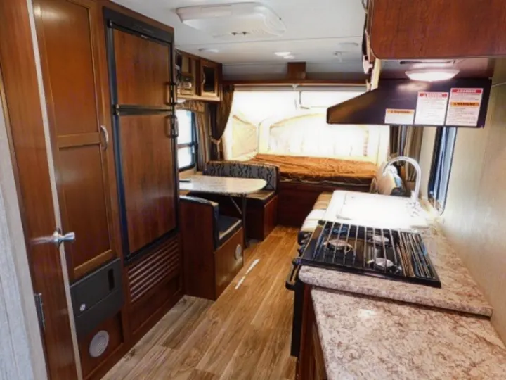 This RV rental interior showcases a compact kitchen with a stove, sink, and counter, a dinette, fridge, and a comfortable bed.. Keystone RV Passport Express 2016