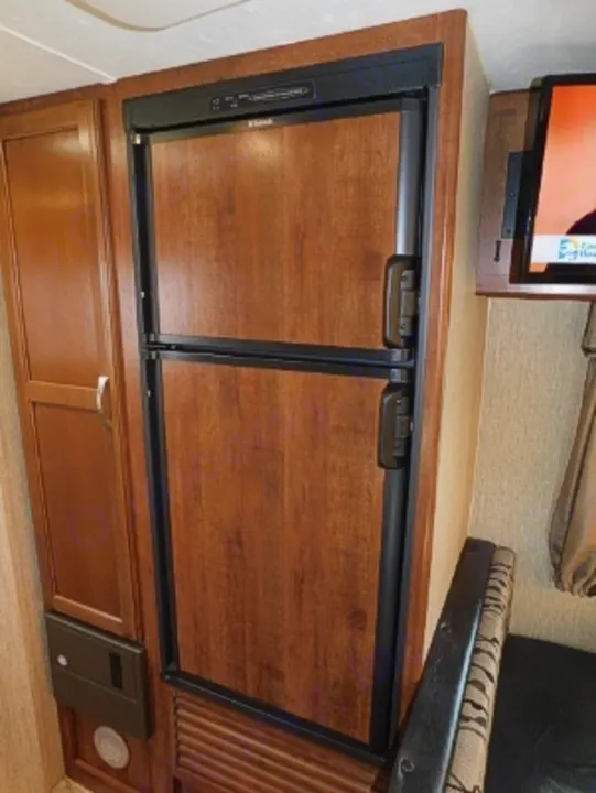 Explore this RV rental's compact kitchen, featuring a two-door wood-paneled refrigerator/freezer, cabinetry, and a wall-mounted TV.. Keystone RV Passport Express 2016