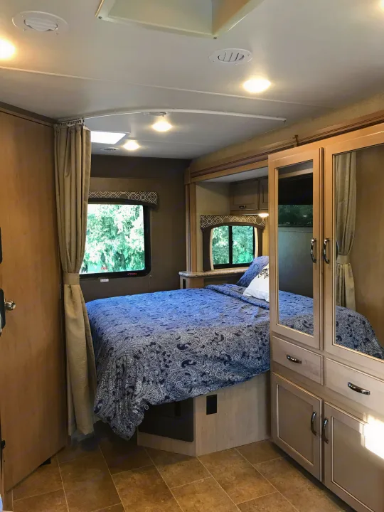 Explore this cozy RV rental bedroom featuring a blue bed, mirrored closet, and bright windows. Perfect for travel.. Thor Chateau 2019
