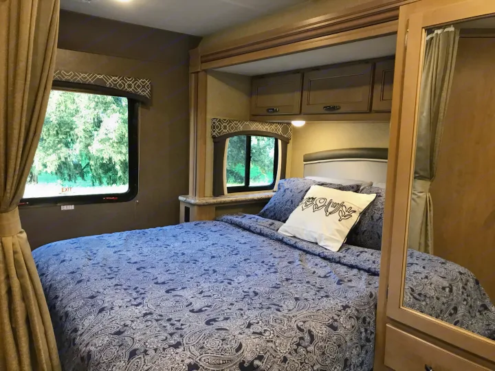 Comfortable RV rental bedroom with blue paisley bedding, windows overlooking nature, and storage cabinets.. Thor Chateau 2019