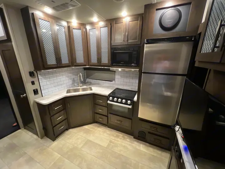 Modern RV rental kitchen features dark cabinets, stainless steel appliances, an L-shaped counter, and subway tile.. Grand Design Momentum 2022