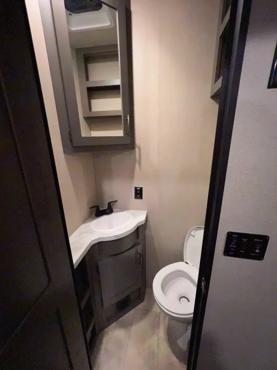 Compact RV rental bathroom with white sink, dark faucet, mirrored medicine cabinet, toilet, & wall controls.. Grand Design Momentum 2022