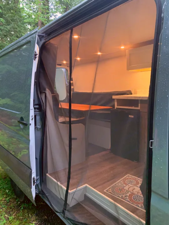 RV rental with open side door and mosquito net, revealing a cozy camper van interior: bed, table, fridge, & "Welcome" mat.. Mercedes-Benz B class campervan MB 140 2014