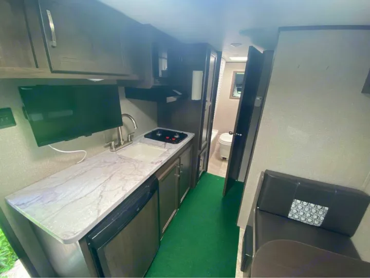 RV rental interior: kitchen with TV, green floor, dinette seating, and bathroom view. Compact comfort for travelers.. Jayco Jay Flight SLX Baja Edition 2020