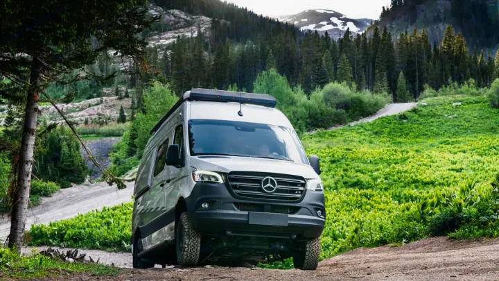 A light grey Mercedes Sprinter RV rental, equipped for off-road, on a dirt path through green mountains.. Winnebago Revel 2022