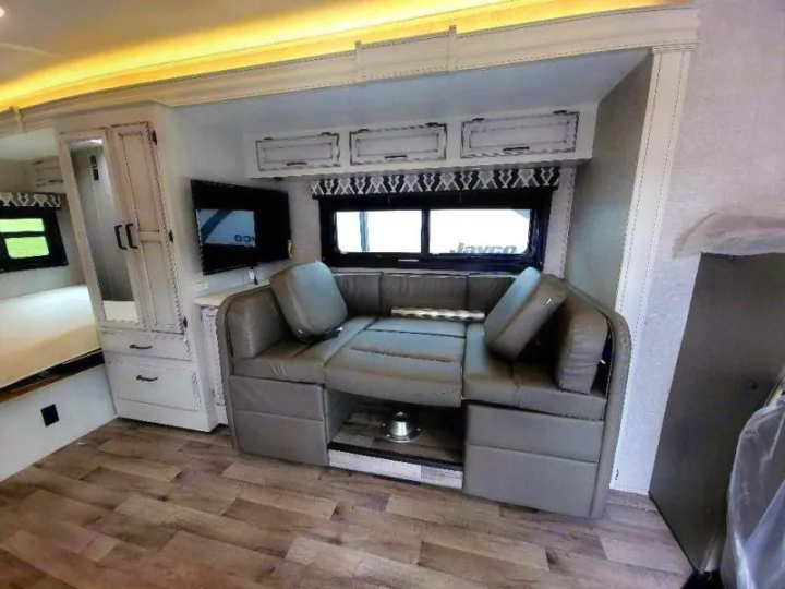 Modern RV rental interior: U-shaped dinette with convertible table, TV, storage, and visible bed area.. Jayco Redhawk 2022